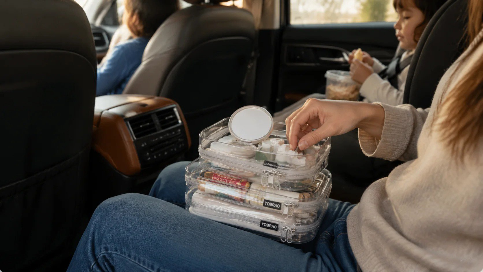 Mother using a small clear hard-shell organizer pouch to access travel toiletries while children ride in car seats in the background of a family SUV.